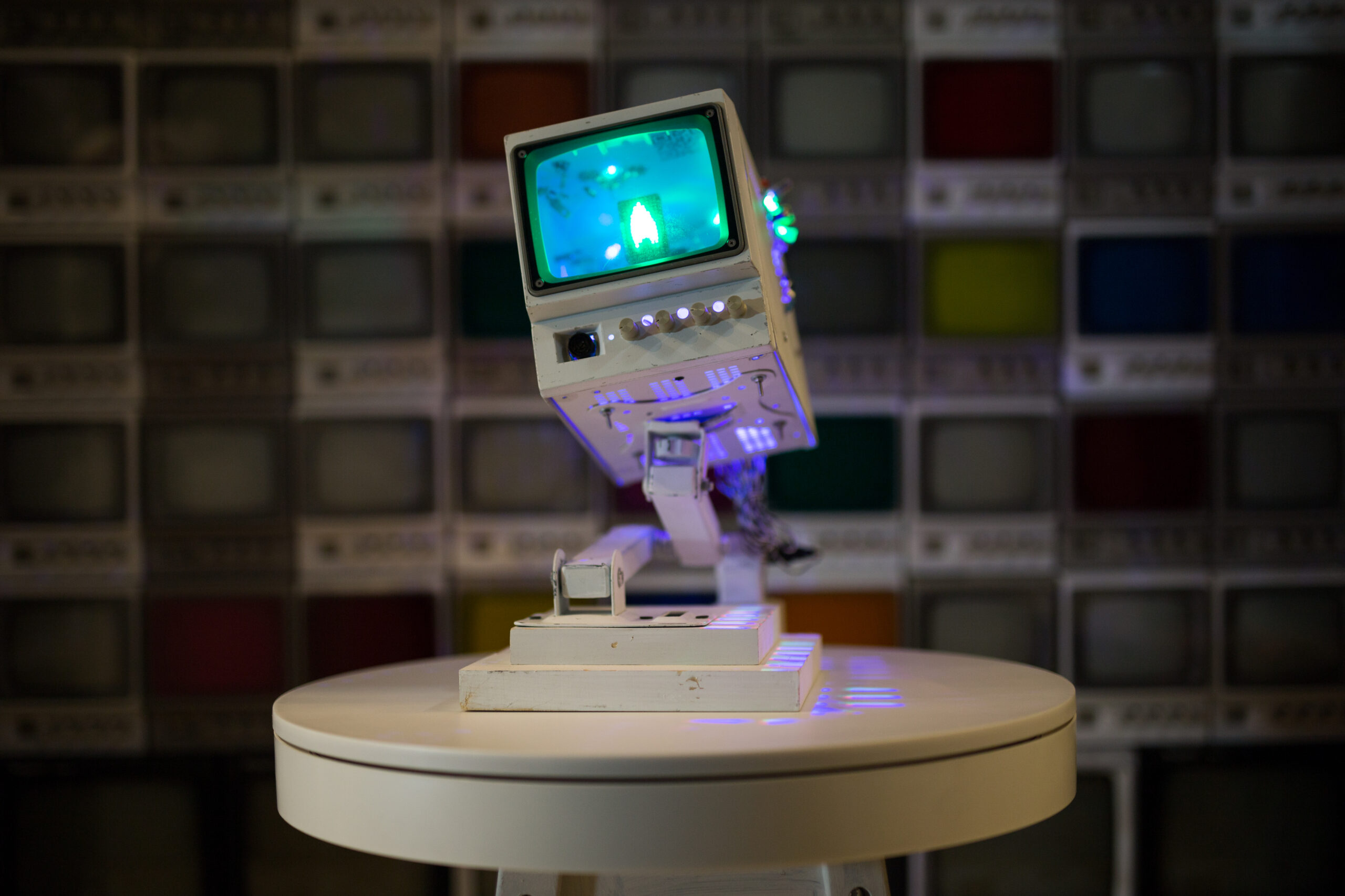 A single white CRT television on a small white table with a candle flame on its screen and a stack of shut-down televisions behind it.
