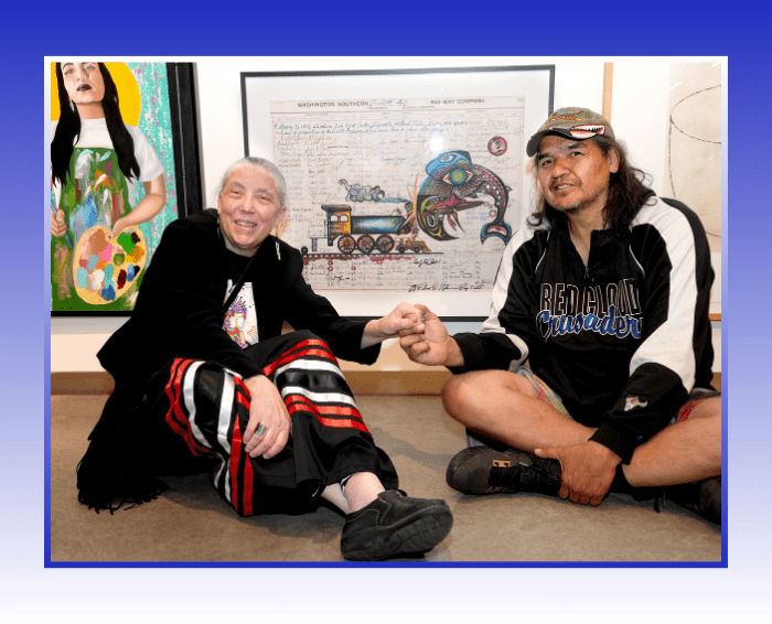 Two people sit holding hands in front of artwork in gallery