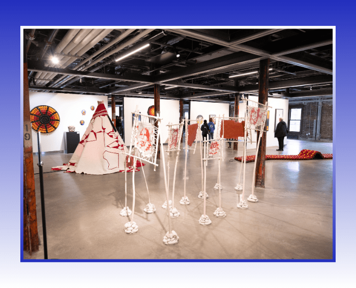 Artwork in Gallery. Red and white teepee and standing white posts with red images of faces on the top