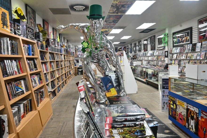 Wide-angle photo of the interior of a comic book store. In the center of the frame is a Silver Surfer statue decorated with St. Patrick's Day merchandise. Surrounding the statue are thousands of comic books on shelves, in boxes and facing outward for display.