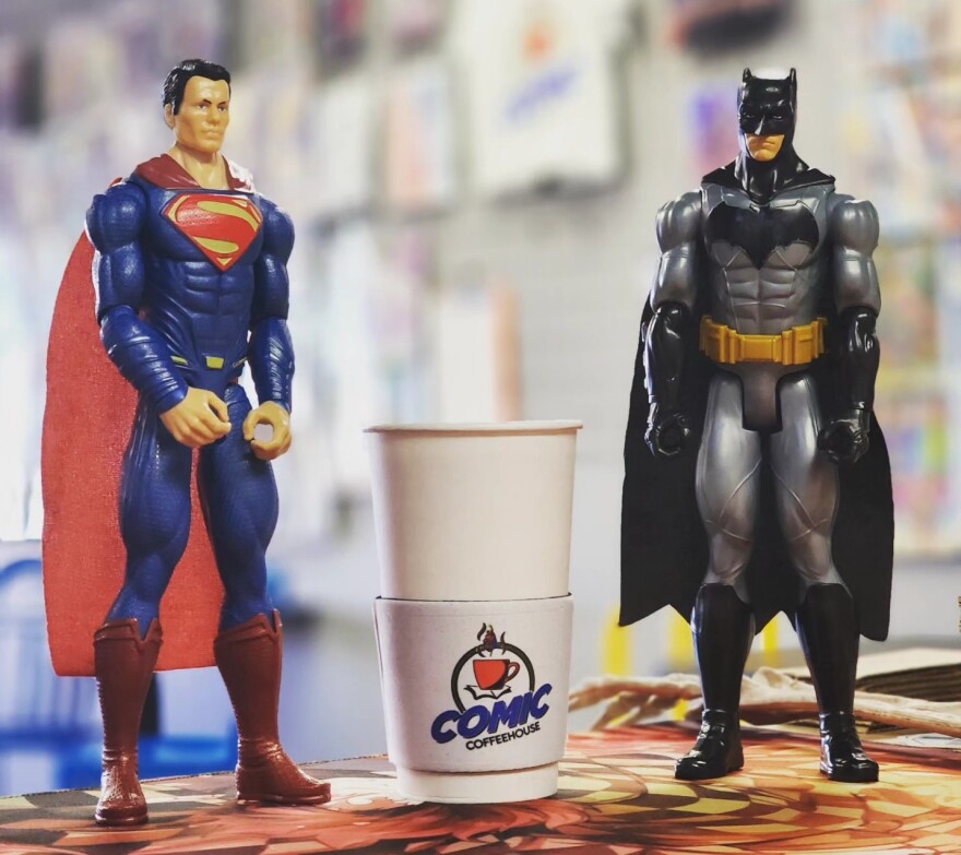 A paper coffee cup with the cardboard sleeve for Comic Coffeehouse is flanked by a Supermand action figure and a Batman action figure.