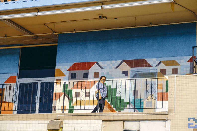 A side of a building has paintings of homes on a wall beyond a wrought iron railing. A person is walking along the landing.