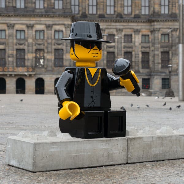 An enormous yellow-and-black Lego figurine, wearing a black hat and black sunglasses, sits on a studded concrete block in the middle of a city square.