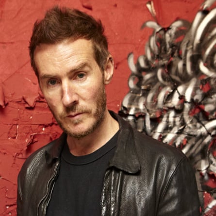 Robert Del Naja, wearing a leather jacket over a T-shirt and with a slight beard and short hair pushed into a peak, stands next to a textured wall