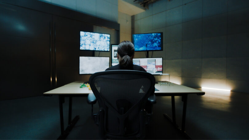 Digital artist Kaoru Tanaka in her studio with her back to viewers, seated in front of her desk and five external monitors and laptop