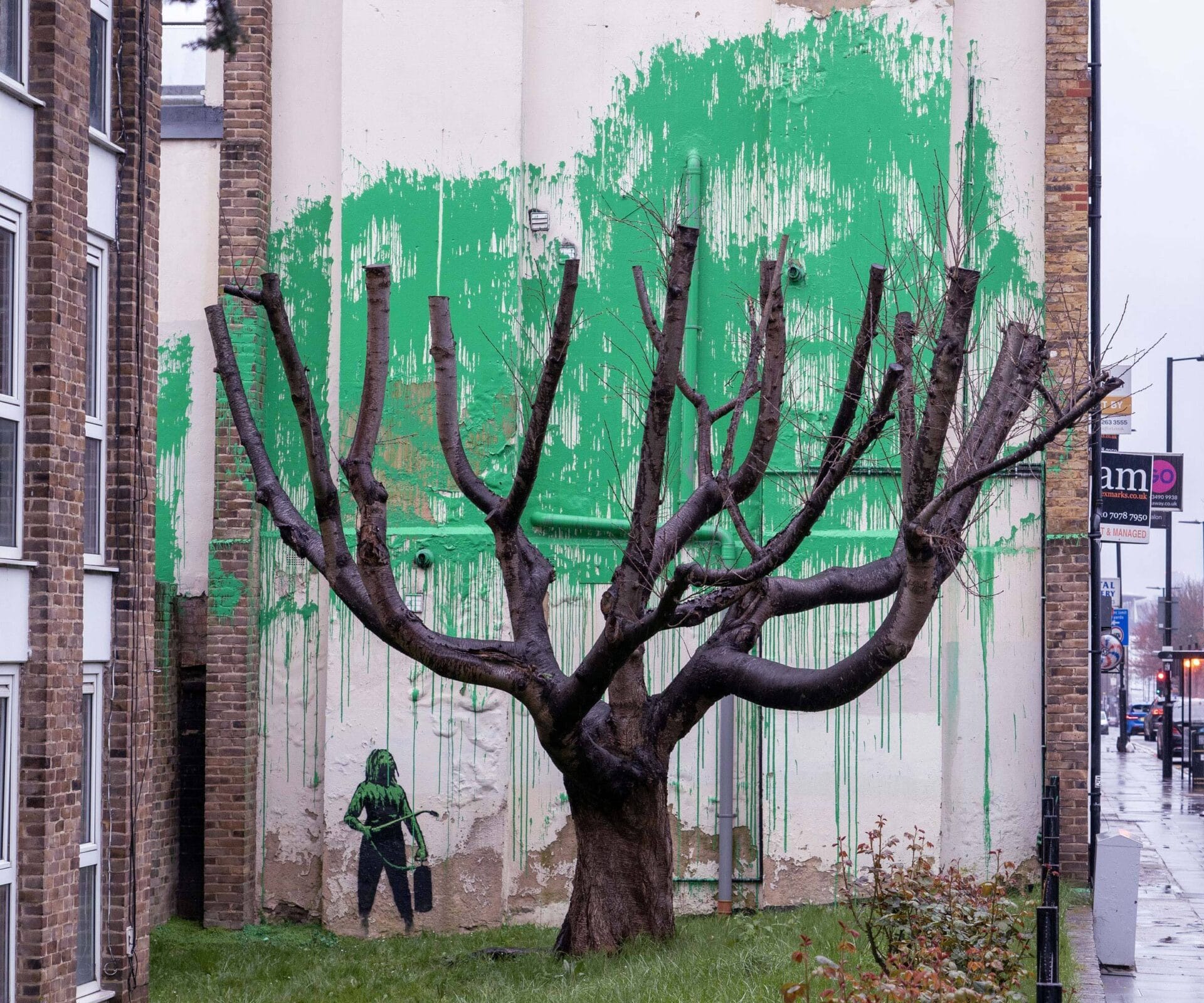 A street art mural behind a recently pollarded tree in London featuring a small figure holding up a paint sprayer and a large swath of green paint that hovers behind the tree like foliage.
