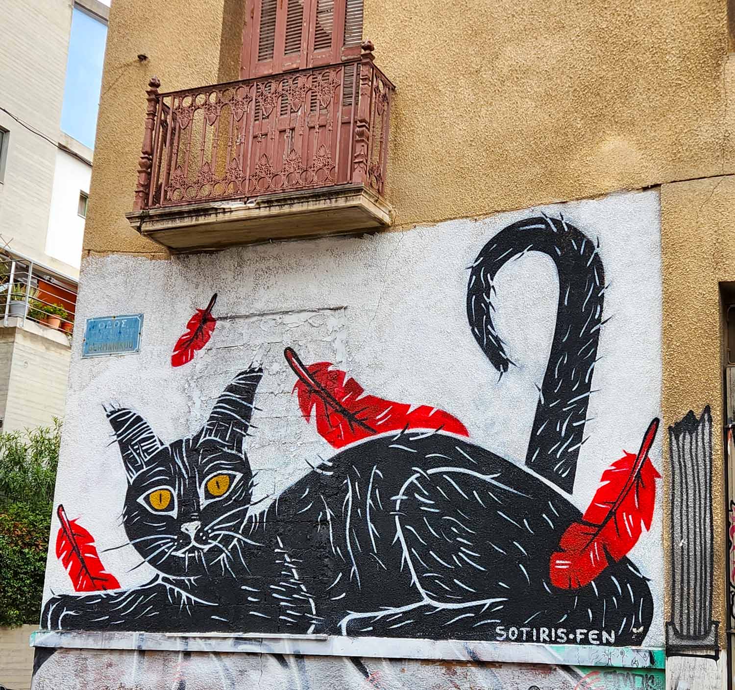 Street art of a black cat with yellow eyes and red feathers falling on a wall in Athens under an old-fashioned balcony - my street art tour of Athens review