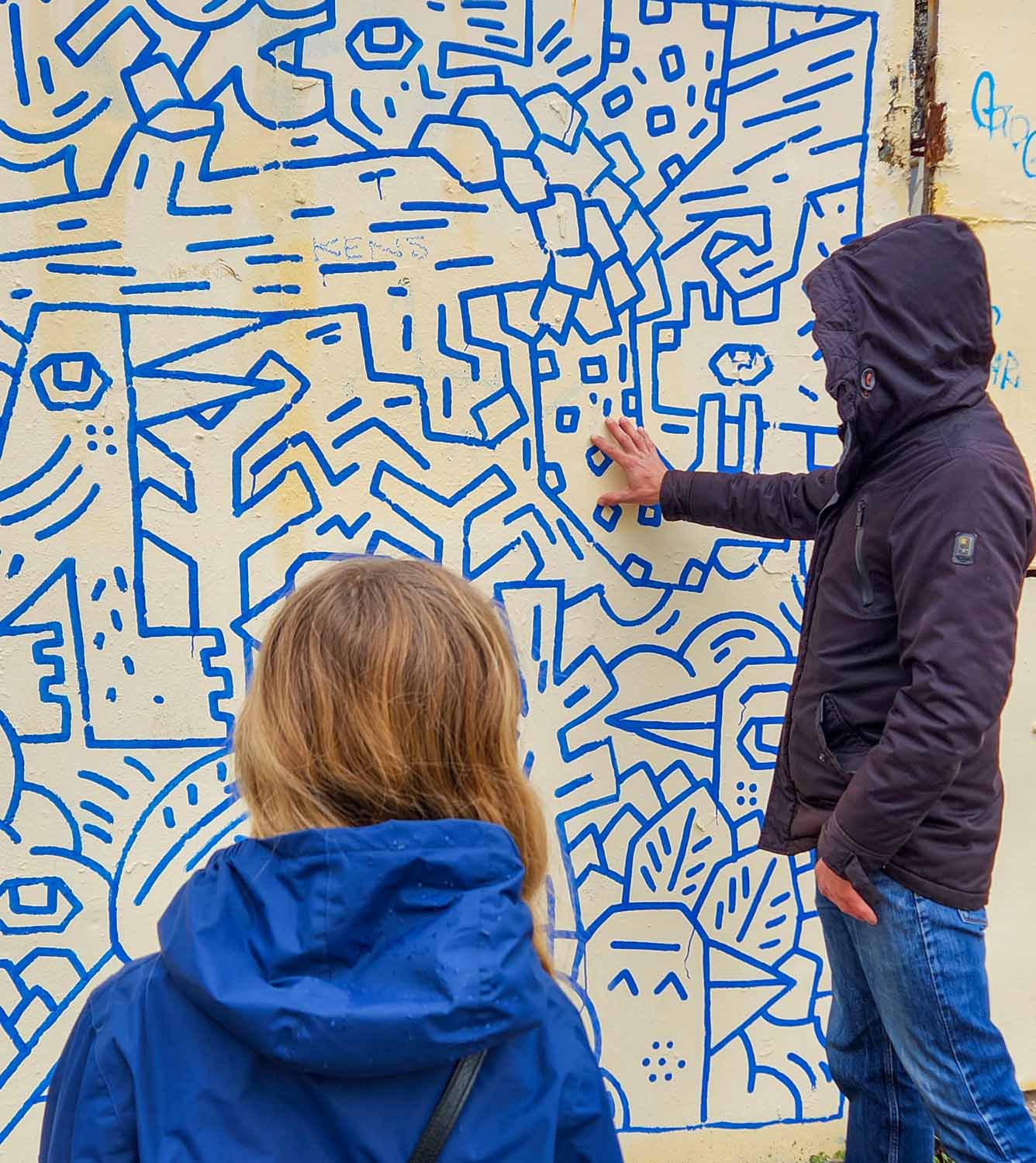 My daughter watches our tour guide Nikos pointing to an artwork made of blue patterns on the cream wall, in a similar style to Keith Haring - my review of a street art tour of Athens with kids