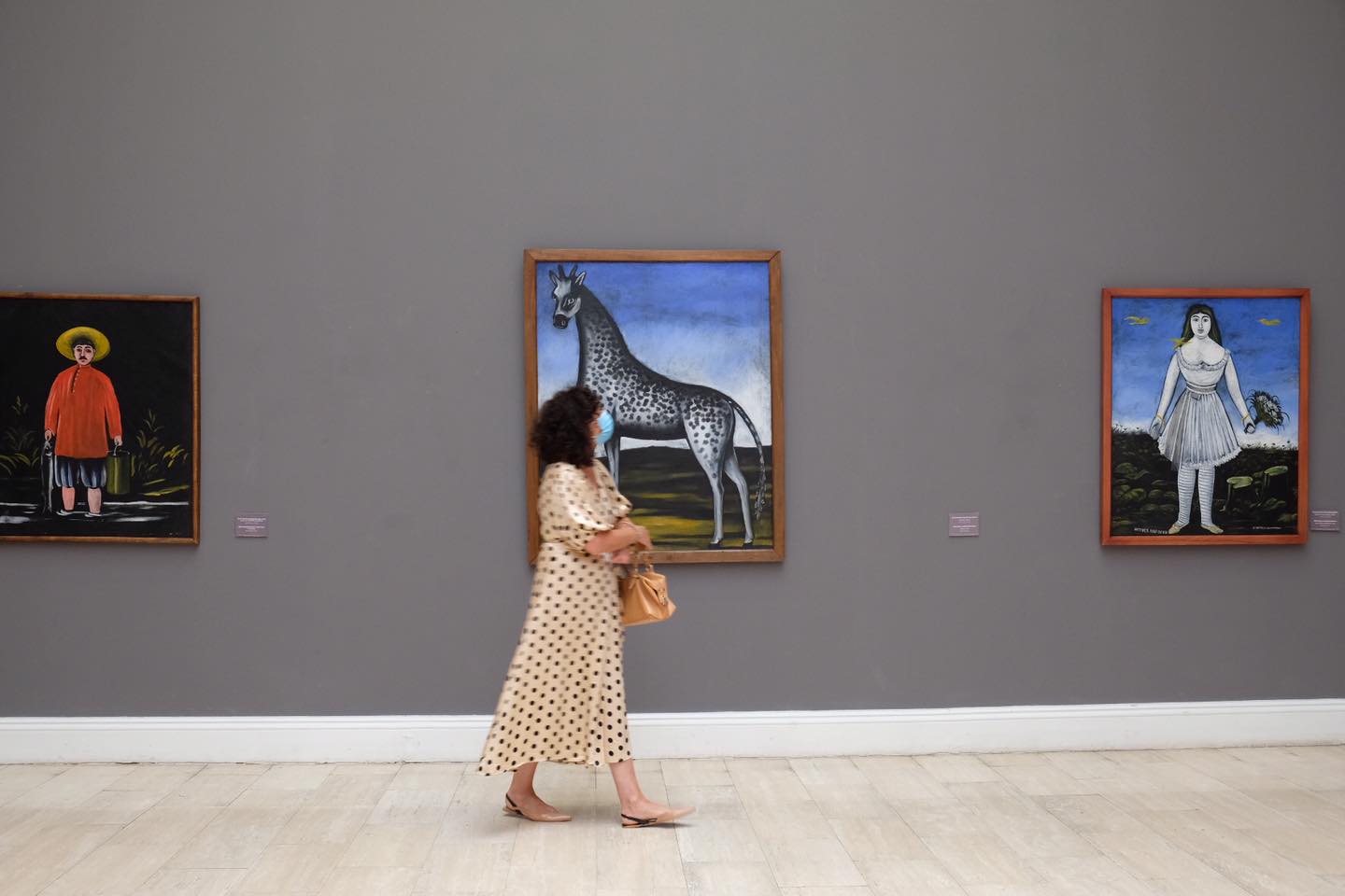 The interior of an art gallery with gray walls; a woman walks past a painting of a giraffe